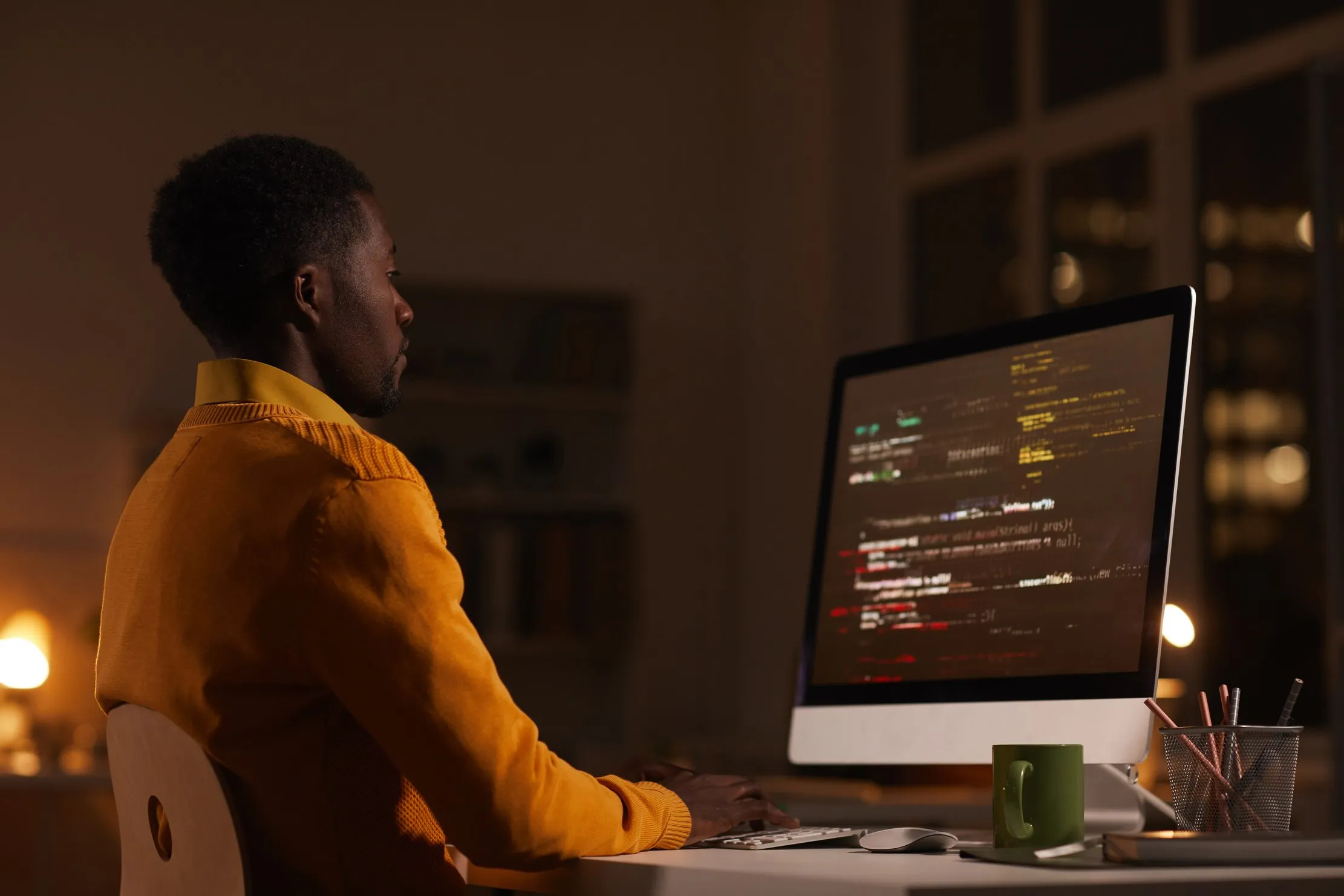 A person working late at a desktop computer showing lines of code in a dim office