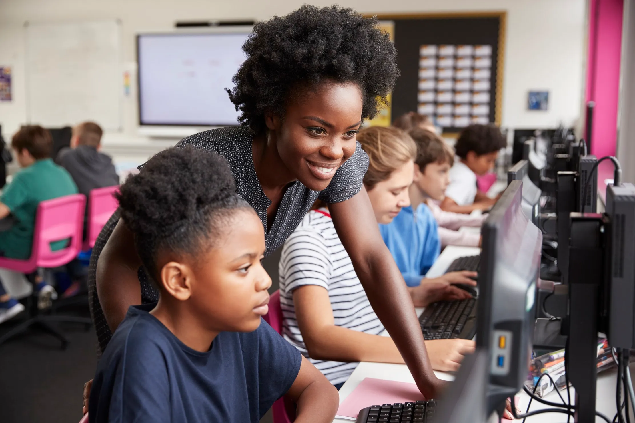 Teacher helping a pupil at a computer in a line of students working at screens, representing a school-management software demonstration