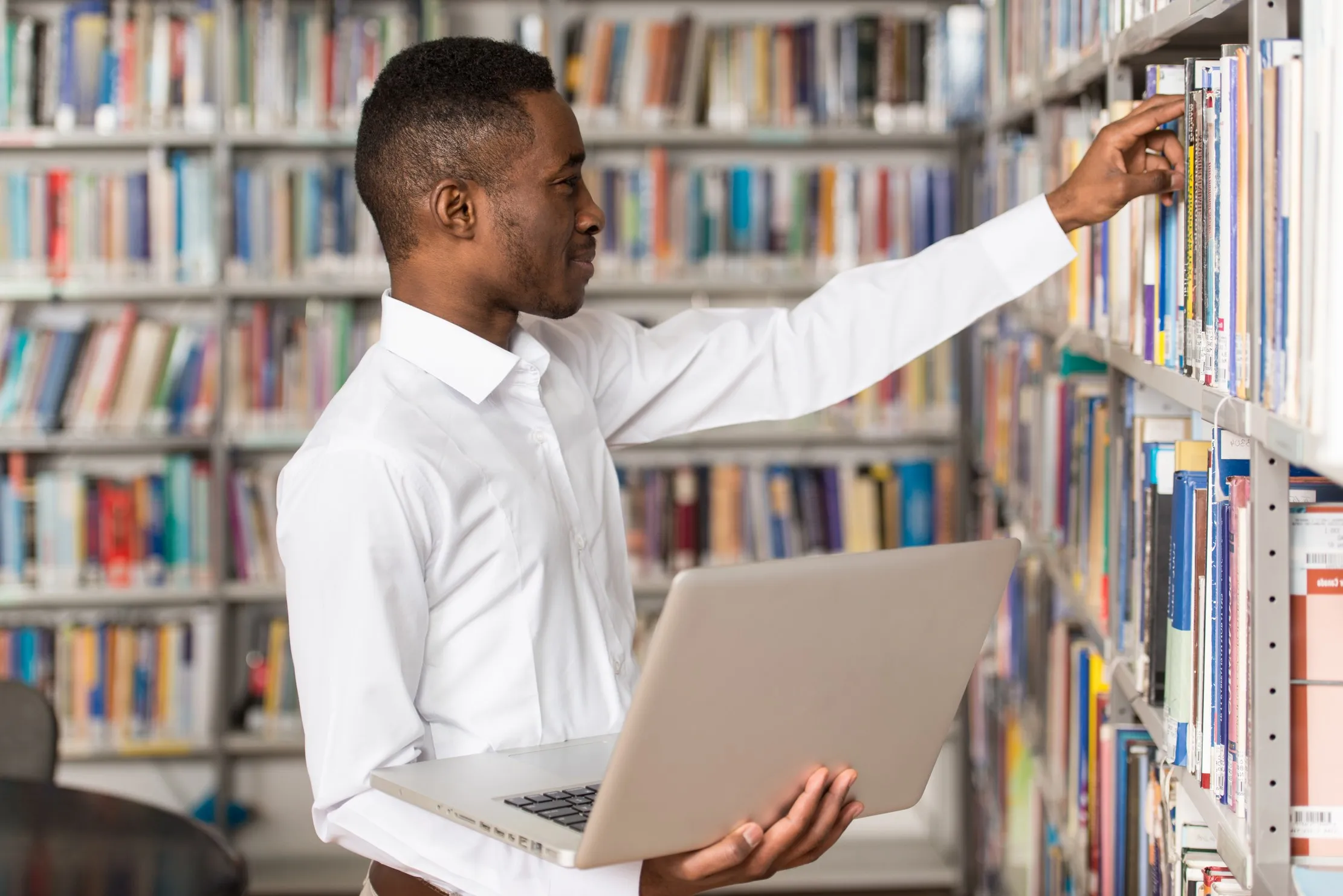 Student reaching for a book in a library while holding a laptop, representing a fictional learner record used to test school software