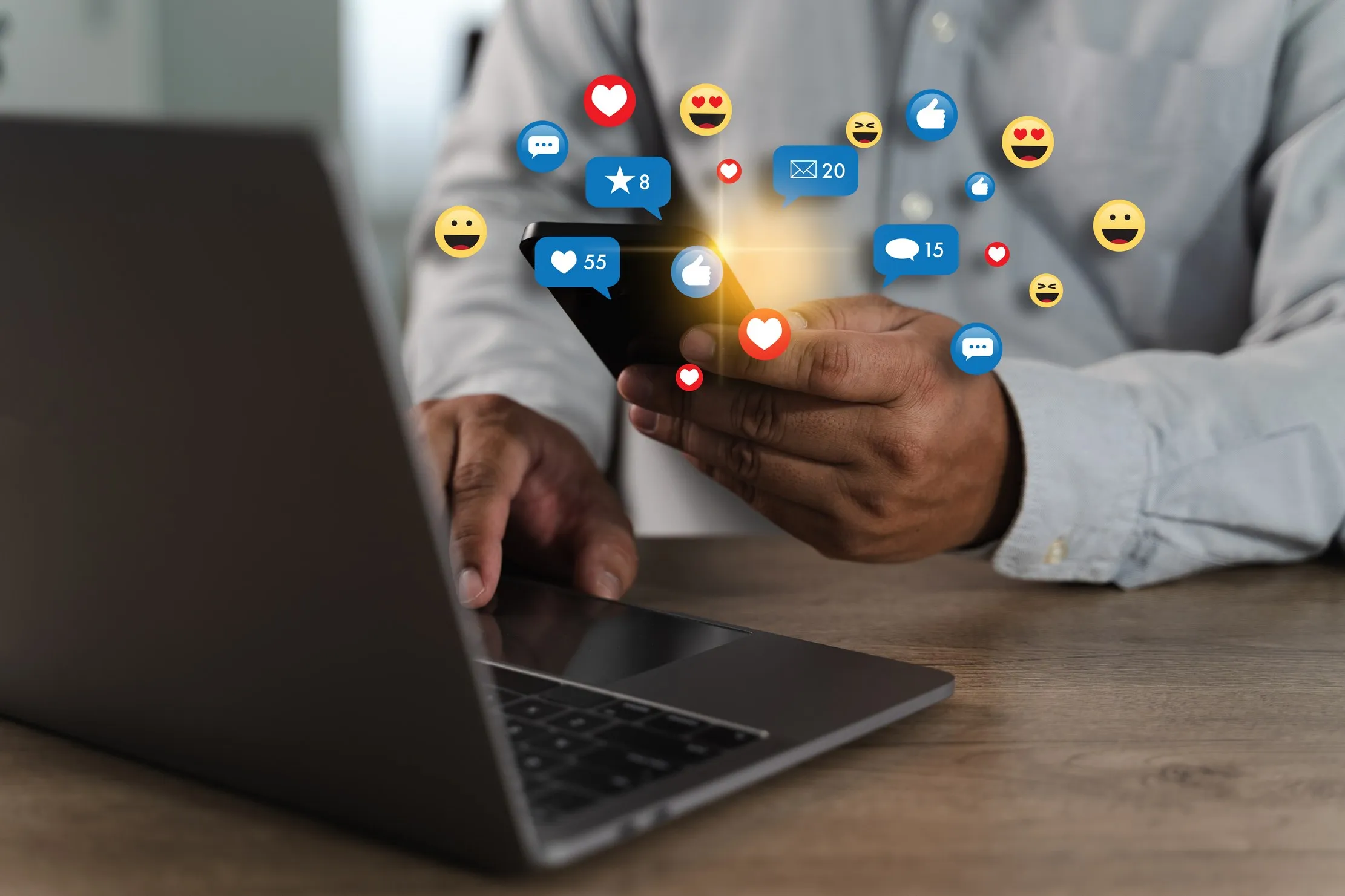 A person using a smartphone beside a laptop with social media reaction icons above the screen