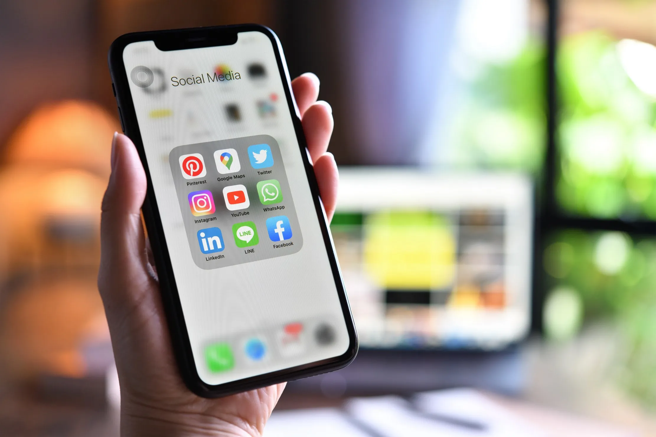 A person holding a smartphone showing a folder of social media app icons, with a laptop blurred behind